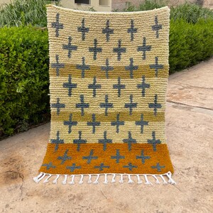 May include: A rectangular rug with a cream-colored background and a repeating pattern of dark blue crosses. The rug has horizontal stripes in shades of yellow and orange, with a fringe along the bottom edge. The rug is made of a thick, textured material.