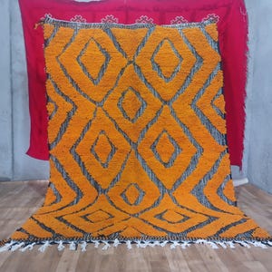 May include: A large, rectangular rug with a vibrant orange diamond pattern on a black background. The rug has a shaggy texture and a white fringe along the bottom edge. The rug is displayed against a red backdrop.