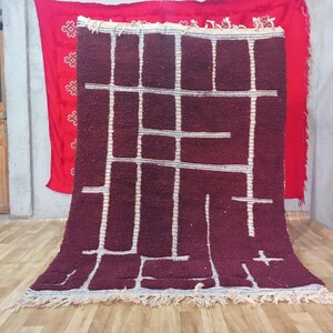 May include: A burgundy rug with a geometric white design. The rug has a rectangular shape with a fringe along the bottom edge. A red textile with white patterns hangs in the background.