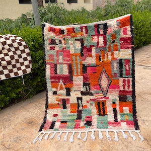 May include: A colorful, rectangular rug with a geometric design. The rug features a mix of orange, pink, green, and black shapes on a cream background. White fringe accents the bottom edge. A smaller, brown and white checkered rug is visible in the background.