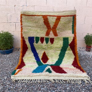 May include: A colorful, hand-woven rug with a geometric design. The rug features a cream-colored background with bold accents in orange, red, blue, and green. The rug has a fringed edge and is displayed outdoors.