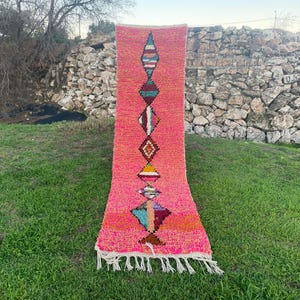 Op de afbeelding: Een felroze tapijt met een geometrisch patroon in verschillende kleuren. Het tapijt heeft ruit- en driehoekvormen in rood, blauw, geel en bruin. Het heeft een gefranjerde rand en wordt op groen gras getoond.