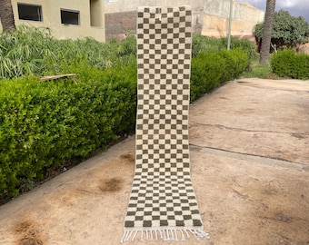May include: A long, rectangular rug with a checkered pattern in shades of beige and brown. The rug has a fringe at the bottom and is displayed outdoors. The rug's design is a classic, geometric pattern.