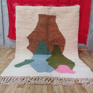 May include: A cream-colored, rectangular rug with a brown, green, blue, and pink abstract design. The rug has a fringe along the bottom edge. The design features shapes resembling vases or organic forms.