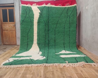 May include: A large, rectangular green rug with a white and black design. The rug has a red border at the top and a fringe along the bottom edge. The rug is displayed on a wooden floor.