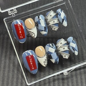May include: A set of artificial nails in a clear plastic case. The nails feature various designs, including a blue base with the word "BUCKIN" in red, a nude and white design with silver accents, a blue and white geometric pattern, and silver bead accents.