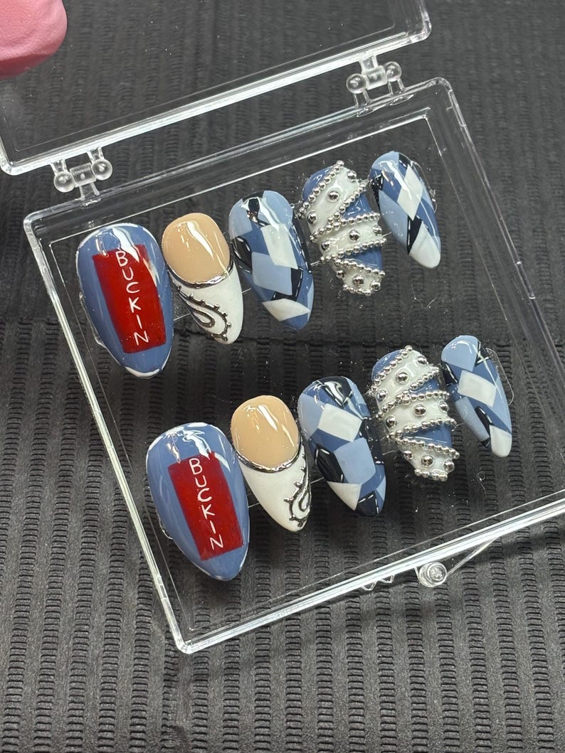 May include: A set of artificial nails in a clear plastic case. The nails feature various designs, including a blue base with the word "BUCKIN" in red, a nude and white design with silver accents, a blue and white geometric pattern, and silver bead accents.