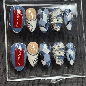 May include: A set of ten artificial nails in a clear plastic case. The nails feature various designs, including blue and white geometric patterns, silver bead accents, and the word "BUCKIN" in red on a blue background. The nails are oval-shaped.