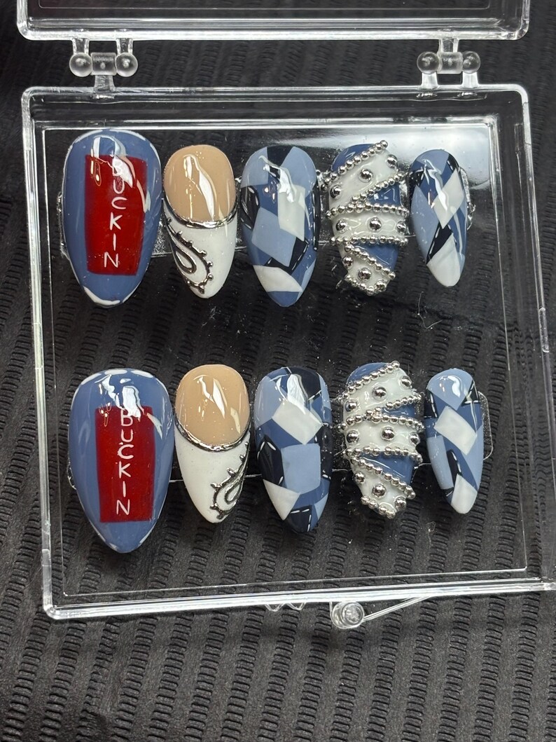 May include: A set of ten artificial nails in a clear plastic case. The nails feature various designs, including blue and white geometric patterns, silver bead accents, and the word "BUCKIN" in red on a blue background. The nails are oval-shaped.