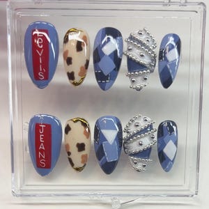 Puede incluir: Un conjunto de u&ntilde;as postizas ovaladas en una caja de pl&aacute;stico transparente. Los dise&ntilde;os incluyen u&ntilde;as azules con texto "JEANS", estampado de vaca, patrones de rombos y adornos blancos y plateados. Presentado en una caja de pl&aacute;stico transparente.