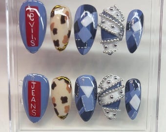 May include: A set of press-on nails in a clear plastic case. The nails feature various designs, including blue with "JEANS" text, cow print, argyle patterns, and white and silver embellishments. The nails are oval-shaped.