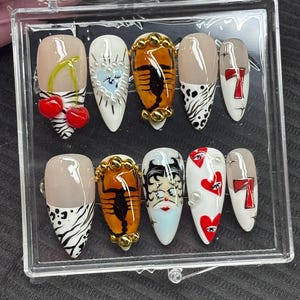 May include: A set of ten artificial nails in a clear plastic case. The nails feature various designs, including cherries, hearts, a scorpion, and Betty Boop. Colors include red, white, black, gold, and nude.