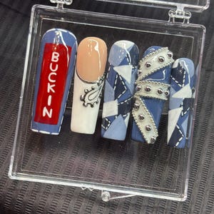 May include: A collection of five artificial nails displayed in a clear case. The nails showcase diverse designs, including a red nail with the word "BUCKIN", a nude nail with a silver paisley, and nails with blue denim patterns and silver embellishments.
