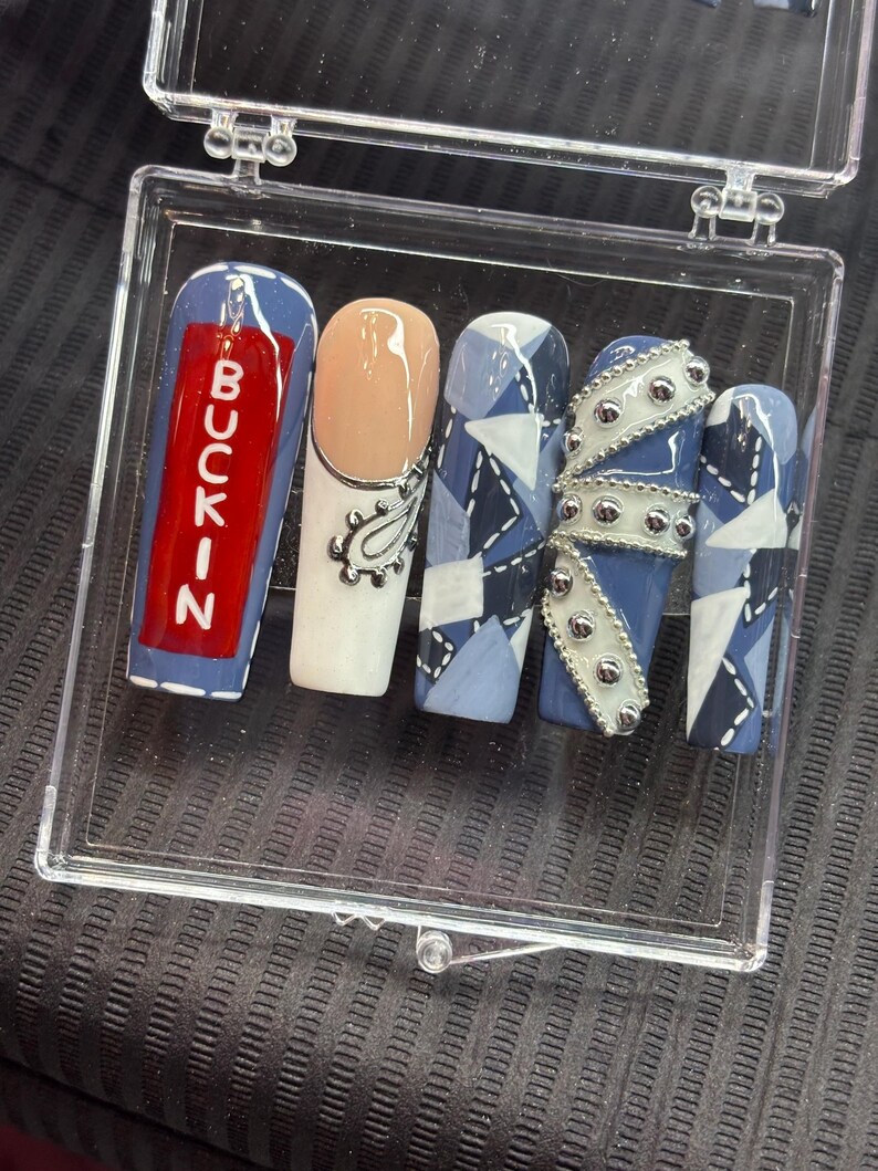 May include: A collection of five artificial nails displayed in a clear case. The nails showcase diverse designs, including a red nail with the word "BUCKIN", a nude nail with a silver paisley, and nails with blue denim patterns and silver embellishments.