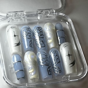 Puede incluir: Un juego de diez uñas postizas azules y blancas con un diseño de mariposa. Las uñas están en una caja de plástico transparente.