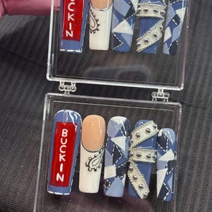 May include: A set of acrylic nails in a clear case. The nails feature various designs, including a red rectangle with the word "BUCKIN" in white, a nude nail with a silver paisley design, and blue and white geometric patterns. Some nails have silver embellishments.