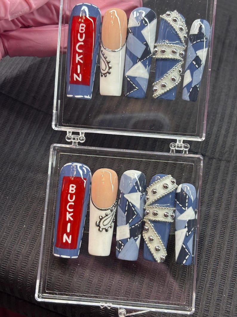 May include: A set of acrylic nails in a clear case. The nails feature various designs, including a red rectangle with the word "BUCKIN" in white, a nude nail with a silver paisley design, and blue and white geometric patterns. Some nails have silver embellishments.