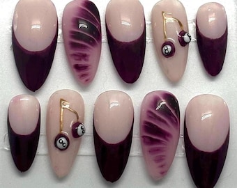 May include: Set of ten false nails with a pink and purple ombre design. Some nails feature a gold cherry charm with a black 8 ball on top.