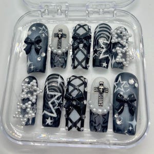 Puede incluir: Un conjunto de diez u&ntilde;as postizas en blanco y negro con varios dise&ntilde;os, incluyendo estrellas, lazos, cruces y patrones a cuadros. Las u&ntilde;as se muestran en una caja de pl&aacute;stico transparente.