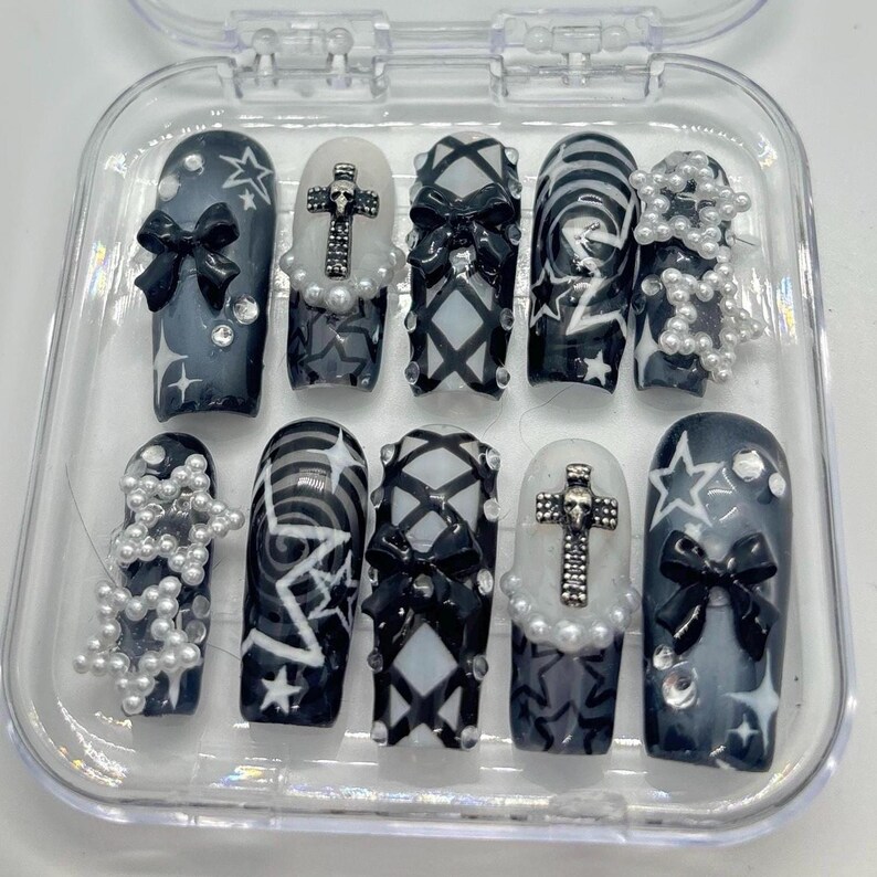 Puede incluir: Un conjunto de diez u&ntilde;as postizas en blanco y negro con varios dise&ntilde;os, incluyendo estrellas, lazos, cruces y patrones a cuadros. Las u&ntilde;as se muestran en una caja de pl&aacute;stico transparente.