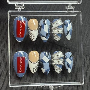 Puede incluir: Un conjunto de uñas postizas en una caja de plástico transparente. Las uñas presentan varios diseños, incluyendo patrones geométricos azules y blancos, detalles de cuentas plateadas y uñas con la palabra "BUCKIN" en letras rojas.