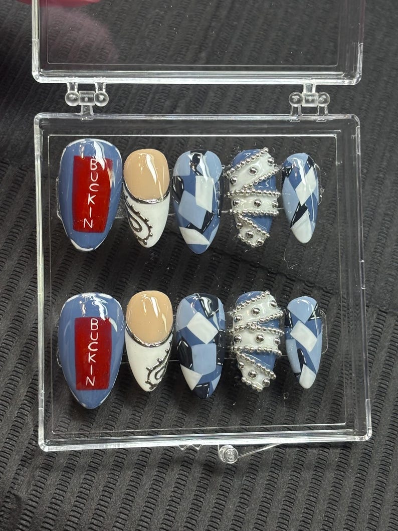May include: A collection of press-on nails displayed in a clear case. The nail designs include blue and white geometric patterns, silver bead embellishments, and nails with the word "BUCKIN" in red. The nails are oval-shaped.