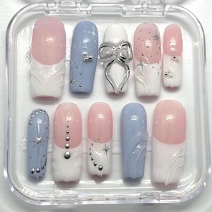 Puede incluir: Un conjunto de diez uñas postizas en un estuche de plástico transparente. Las uñas son una mezcla de azul claro y rosa con puntas blancas. Algunas uñas tienen purpurina plateada, estrellas plateadas y cuentas plateadas. Una uña tiene un lazo plateado.