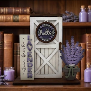 Puede incluir: Un expositor decorativo de madera que incluye una puerta de granero blanca con un cartel redondo de corona que dice hello, un letrero vertical que dice farm fresh LAVENDER y una planta de lavanda en maceta sobre una base de madera.