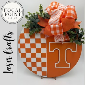 May include: A round wooden sign with a checkerboard pattern and the letter "T" in orange and white. The sign is decorated with a large orange and white bow, greenery, and the text "Tennessee". The sign is supported by a wooden stand.
