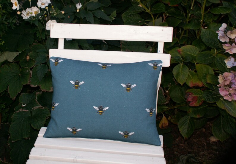 May include: A teal blue throw pillow with a repeating pattern of yellow and black bumblebees. The pillow is sitting on a white wooden bench.