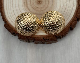 Puede incluir: Dos pendientes redondos dorados con un patrón texturizado en forma de rejilla. Los pendientes se muestran sobre una rodaja de madera con un fondo claro y texturizado.