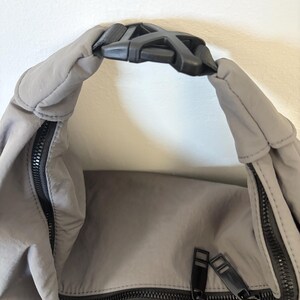 Puede incluir: Un bolso de nailon gris con una hebilla de pl&aacute;stico negra en el asa superior. El bolso incluye un compartimento principal con cremallera negra y un bolsillo frontal con cremallera negra, ambos con tiradores de cremallera rectangulares.