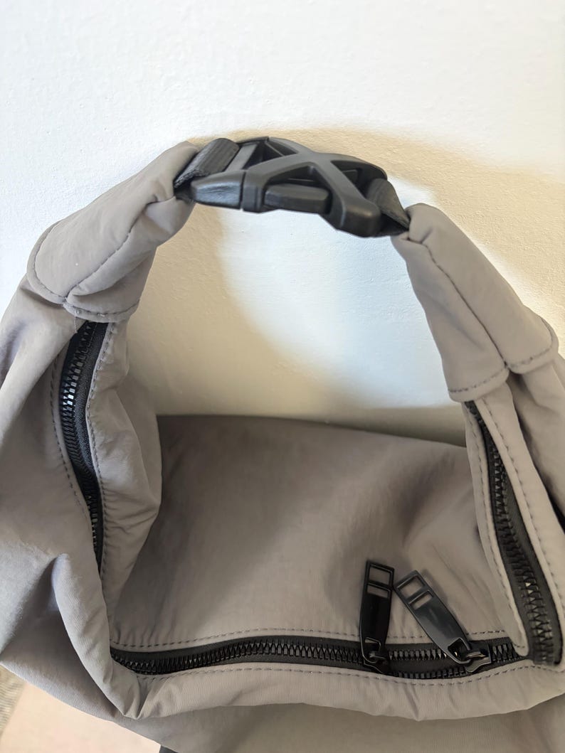 Puede incluir: Un bolso de nailon gris con una hebilla de pl&aacute;stico negra en el asa superior. El bolso incluye un compartimento principal con cremallera negra y un bolsillo frontal con cremallera negra, ambos con tiradores de cremallera rectangulares.