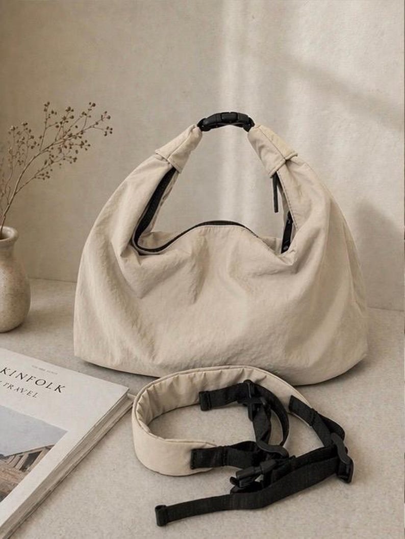 Puede incluir: Un bolso estilo hobo de color beige hecho de una tela ligera y arrugada. El bolso cuenta con una abertura superior asegurada por una hebilla de pl&aacute;stico negra. Se incluye una correa de hombro acolchada a juego con correas de nailon negro.