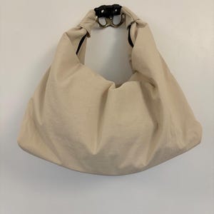 Puede incluir: Un bolso estilo hobo de color beige hecho de una tela lisa de color s&oacute;lido. El bolso presenta una silueta holgada y sin estructura con un asa integrada &uacute;nica. En la parte superior central, el asa est&aacute; asegurada por una hebilla de pl&aacute;stico negro.