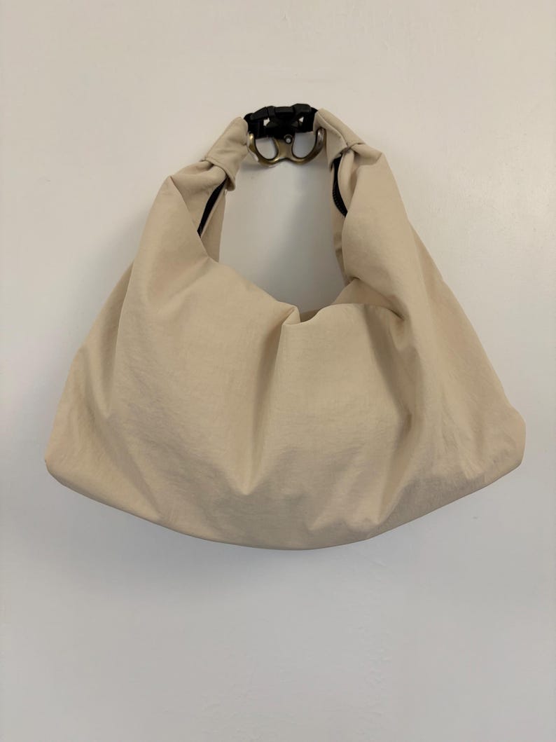 Puede incluir: Un bolso estilo hobo de color beige hecho de una tela lisa de color s&oacute;lido. El bolso presenta una silueta holgada y sin estructura con un asa integrada &uacute;nica. En la parte superior central, el asa est&aacute; asegurada por una hebilla de pl&aacute;stico negro.