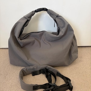 Puede incluir: Un bolso tipo hobo gris con acabado mate y liso, que cuenta con un asa superior asegurada por una hebilla de liberaci&oacute;n lateral de pl&aacute;stico negro. Debajo se encuentra una correa de hombro acolchada gris a juego con nailon negro y herrajes.