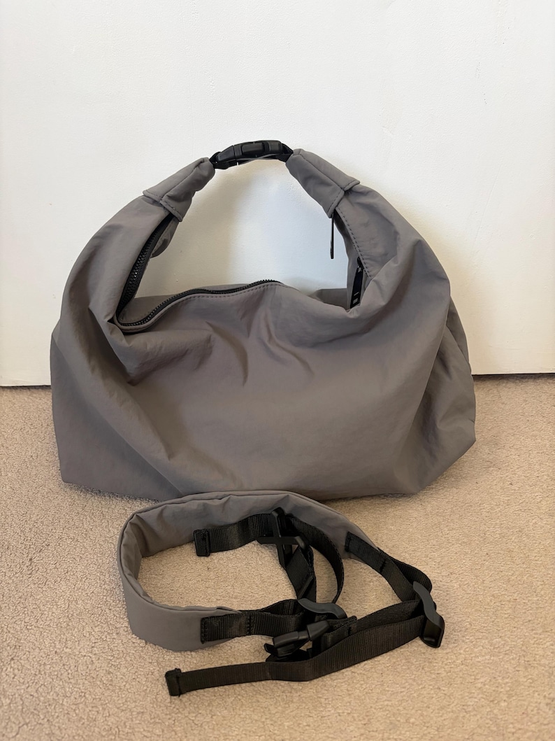 Puede incluir: Un bolso tipo hobo gris con acabado mate y liso, que cuenta con un asa superior asegurada por una hebilla de liberaci&oacute;n lateral de pl&aacute;stico negro. Debajo se encuentra una correa de hombro acolchada gris a juego con nailon negro y herrajes.