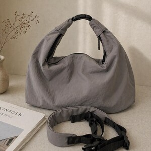 Puede incluir: Un bolso estilo hobo gris hecho de tela arrugada, con un asa superior asegurada con una hebilla de pl&aacute;stico negra. El bolso incluye una correa de nailon negra ajustable y extra&iacute;ble con herrajes de pl&aacute;stico y un cierre de cremallera superior.