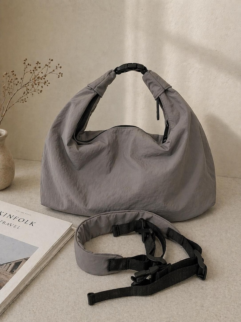 Puede incluir: Un bolso estilo hobo gris hecho de tela arrugada, con un asa superior asegurada con una hebilla de pl&aacute;stico negra. El bolso incluye una correa de nailon negra ajustable y extra&iacute;ble con herrajes de pl&aacute;stico y un cierre de cremallera superior.