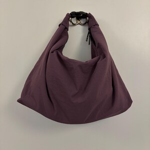 Puede incluir: Un bolso de hombro estilo hobo de color ciruela hecho de una tela suave y ligera. El bolso cuenta con una sola correa integrada que se conecta en la parte superior con una hebilla de liberaci&oacute;n lateral de pl&aacute;stico negro. Silueta holgada.