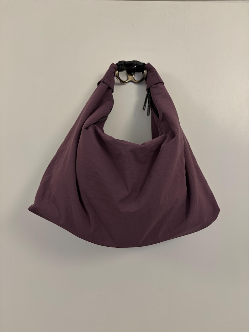 Puede incluir: Un bolso de hombro estilo hobo de color ciruela hecho de una tela suave y ligera. El bolso cuenta con una sola correa integrada que se conecta en la parte superior con una hebilla de liberaci&oacute;n lateral de pl&aacute;stico negro. Silueta holgada.