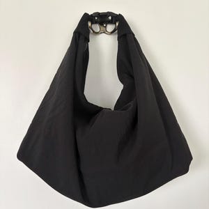 Puede incluir: Un bolso de estilo hobo negro hecho de una tela suave y ligera. El bolso cuenta con una correa de hombro ancha e integrada que se conecta en la parte superior con una hebilla de liberaci&oacute;n lateral de pl&aacute;stico negro y un anillo met&aacute;lico plateado.