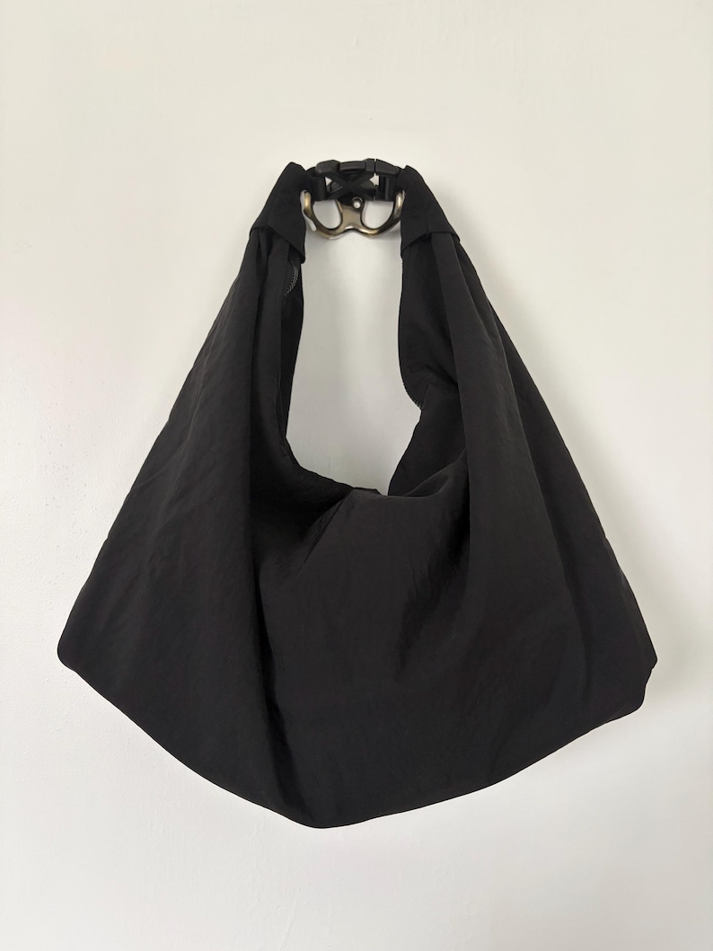 Puede incluir: Un bolso de estilo hobo negro hecho de una tela suave y ligera. El bolso cuenta con una correa de hombro ancha e integrada que se conecta en la parte superior con una hebilla de liberaci&oacute;n lateral de pl&aacute;stico negro y un anillo met&aacute;lico plateado.