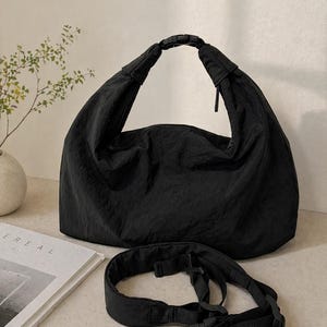 Puede incluir: Un bolso tipo hobo negro de tela arrugada, con un asa superior asegurada por una hebilla de liberaci&oacute;n lateral de pl&aacute;stico negro. Una correa ajustable negra a juego con hebillas de pl&aacute;stico se encuentra frente al bolso.