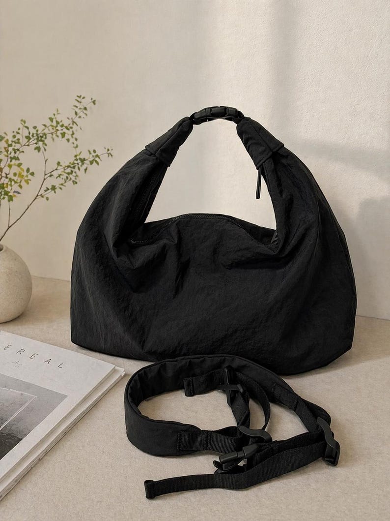 Puede incluir: Un bolso tipo hobo negro de tela arrugada, con un asa superior asegurada por una hebilla de liberaci&oacute;n lateral de pl&aacute;stico negro. Una correa ajustable negra a juego con hebillas de pl&aacute;stico se encuentra frente al bolso.