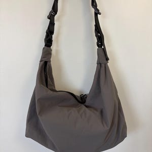 Puede incluir: Un bolso de hombro estilo hobo en color gris carb&oacute;n, confeccionado en tela lisa y mate. El bolso incluye una correa negra ajustable con hebillas de pl&aacute;stico integradas y un cierre de cremallera negro central en la abertura superior.