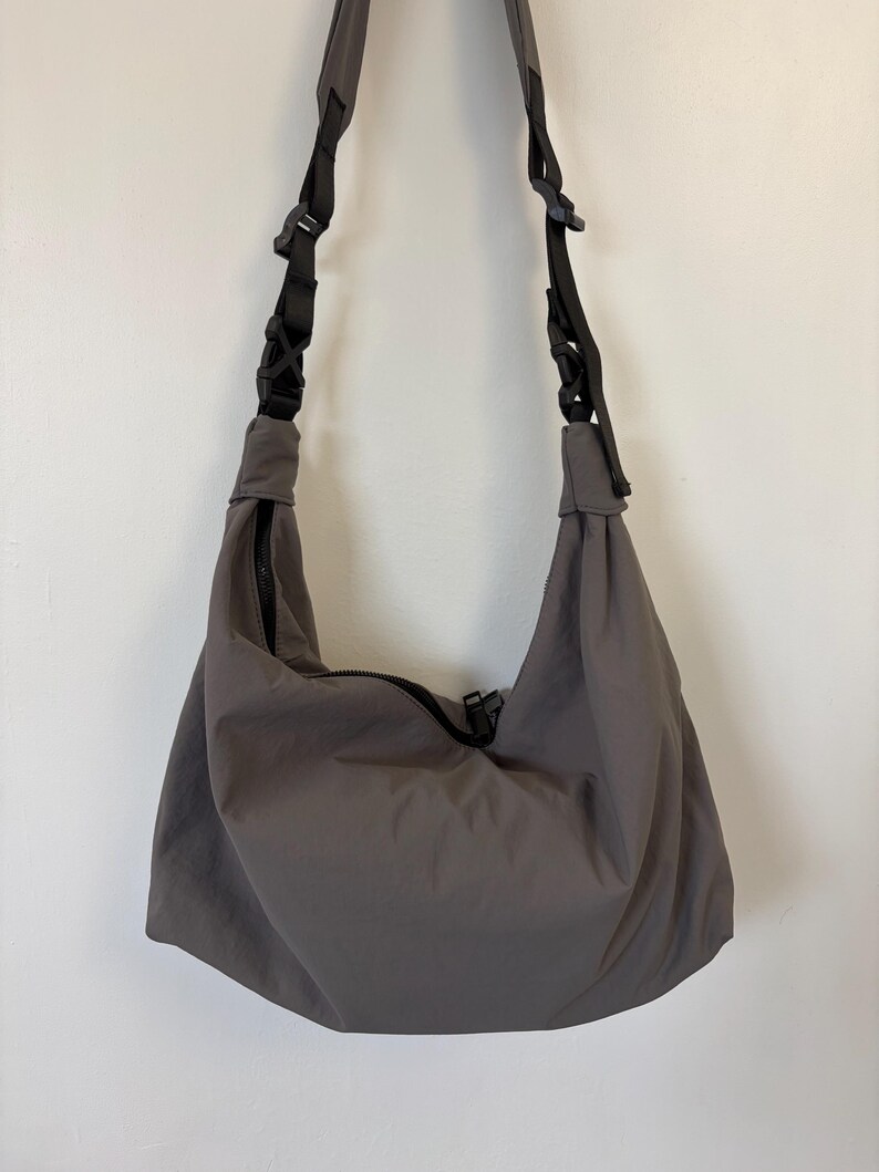 Puede incluir: Un bolso de hombro estilo hobo en color gris carb&oacute;n, confeccionado en tela lisa y mate. El bolso incluye una correa negra ajustable con hebillas de pl&aacute;stico integradas y un cierre de cremallera negro central en la abertura superior.