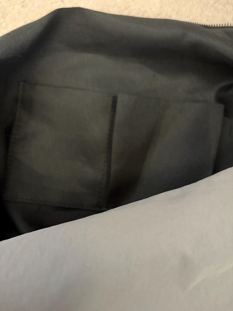 Puede incluir: El interior de un bolso negro con un forro de tela suave y un bolsillo abierto rectangular cosido en el panel posterior. El bolsillo est&aacute; dividido en dos secciones por una costura vertical, proporcionando espacio para guardar objetos peque&ntilde;os.
