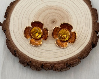 Puede incluir: Un par de pendientes dorados con forma de flor y un gran centro de cristal color ámbar. Los pendientes se exhiben sobre una rodaja de madera con un fondo claro.